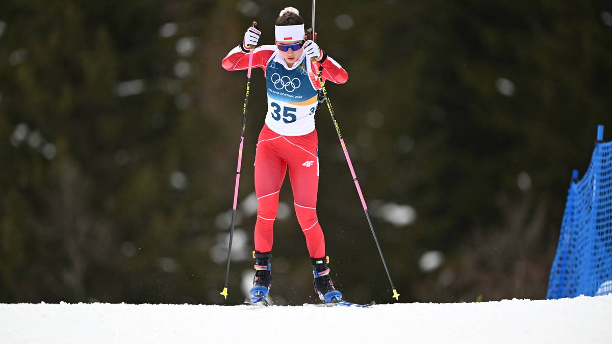 Biathlonistka w czerwonym stroju z numerem 35 na piersi, w pozycji stojącej na śniegu, trzymająca kije narciarskie, z goglami na oczach i białą opaską na głowie.