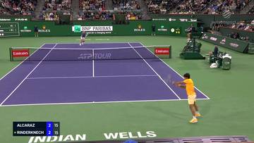 Mecz tenisowy Carlos Alcaraz kontra Arthur Rinderknech na korcie w Indian Wells. Alcaraz w ż&oacute;łtej koszulce serwuje, Rinderknech w białej koszulce i pomarańczowych spodenkach odbiera piłkę. Widoczna jest tablica wynik&oacute;w z aktualnym stanem meczu.