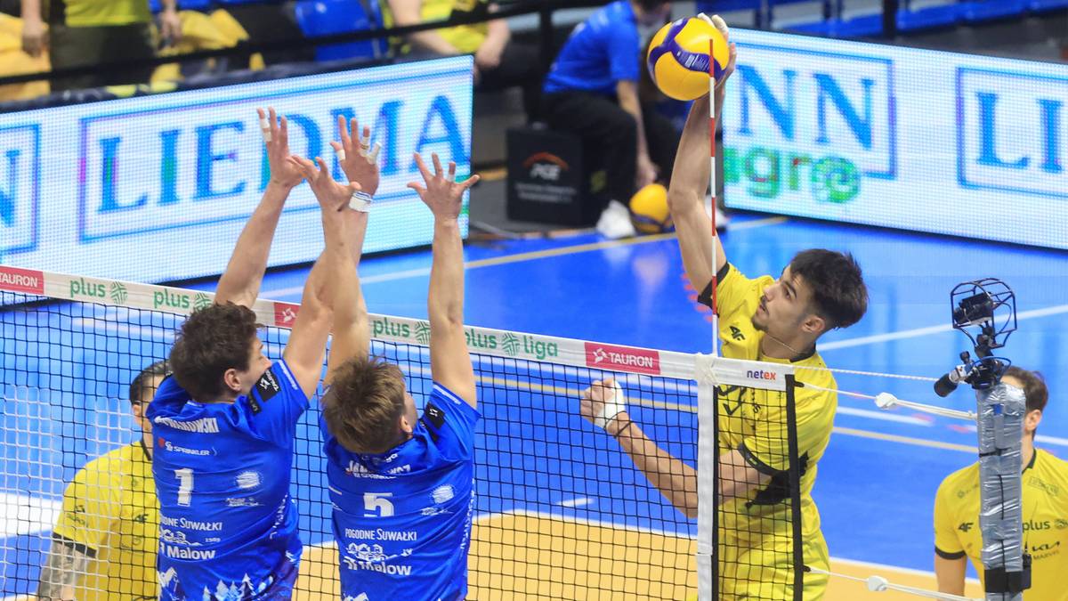 Siatkarz w żółtej koszulce blokuje atak dwóch siatkarzy w niebieskich koszulkach.