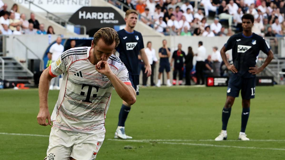 Piłkarz Harry Kane z klubu Bayern Monachium w białej koszulce z czerwonymi pasami świętuje bramkę, trzymając rękę przy ustach. W tle widać innych piłkarzy i trybuny z kibicami.