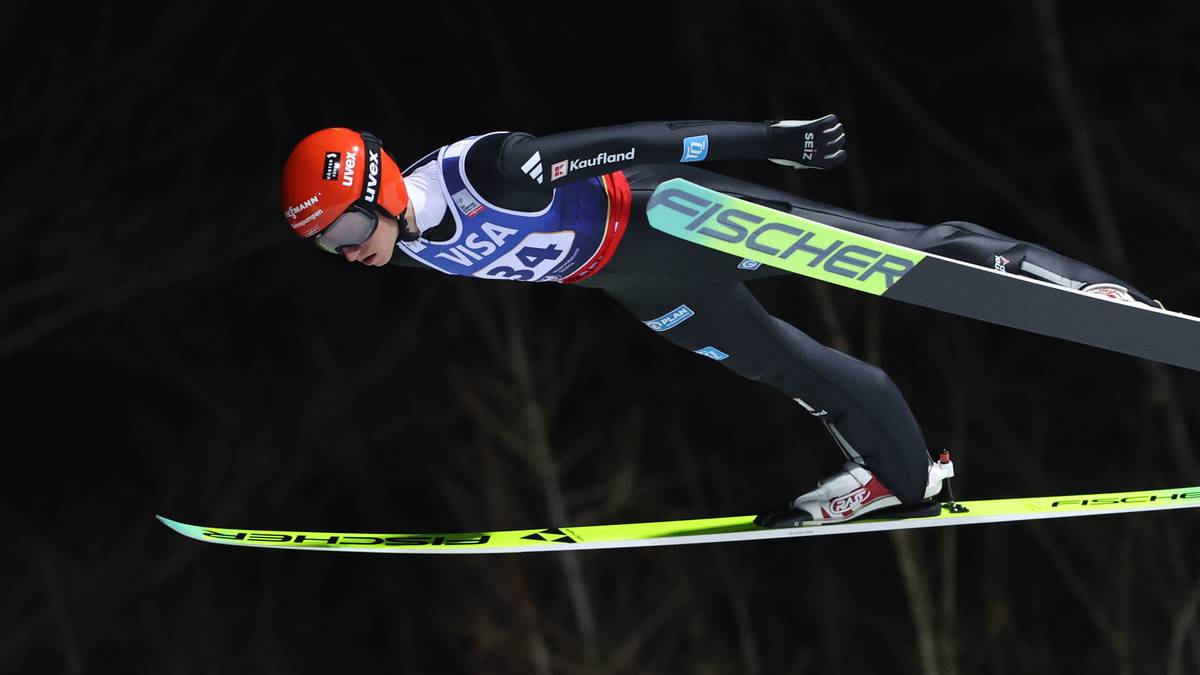 Skoczek narciarski w kombinezonie i kasku z nartami na nogach, podczas lotu.