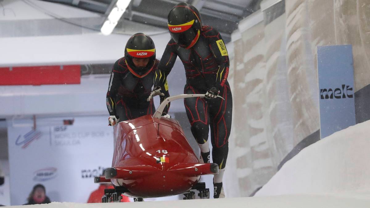 Dwie kobiety w kaskach i kombinezonach do bobslejów pchają czerwone sanie na lodowym torze.