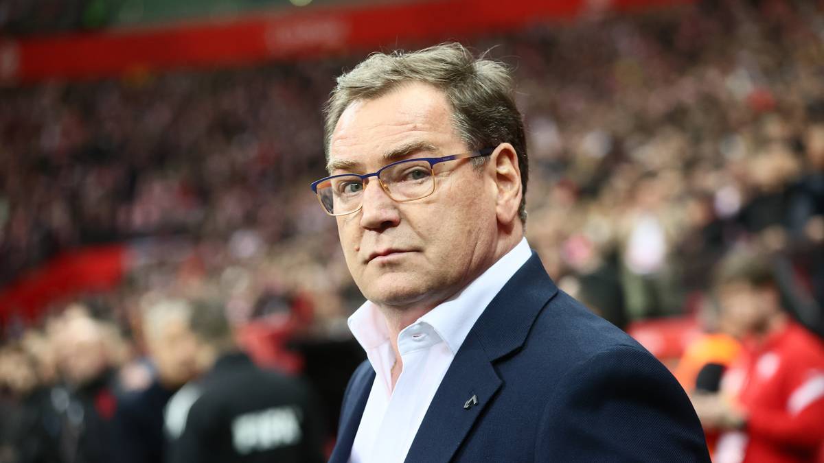 Portret mężczyzny w garniturze i okularach, z widownią stadionu w tle.