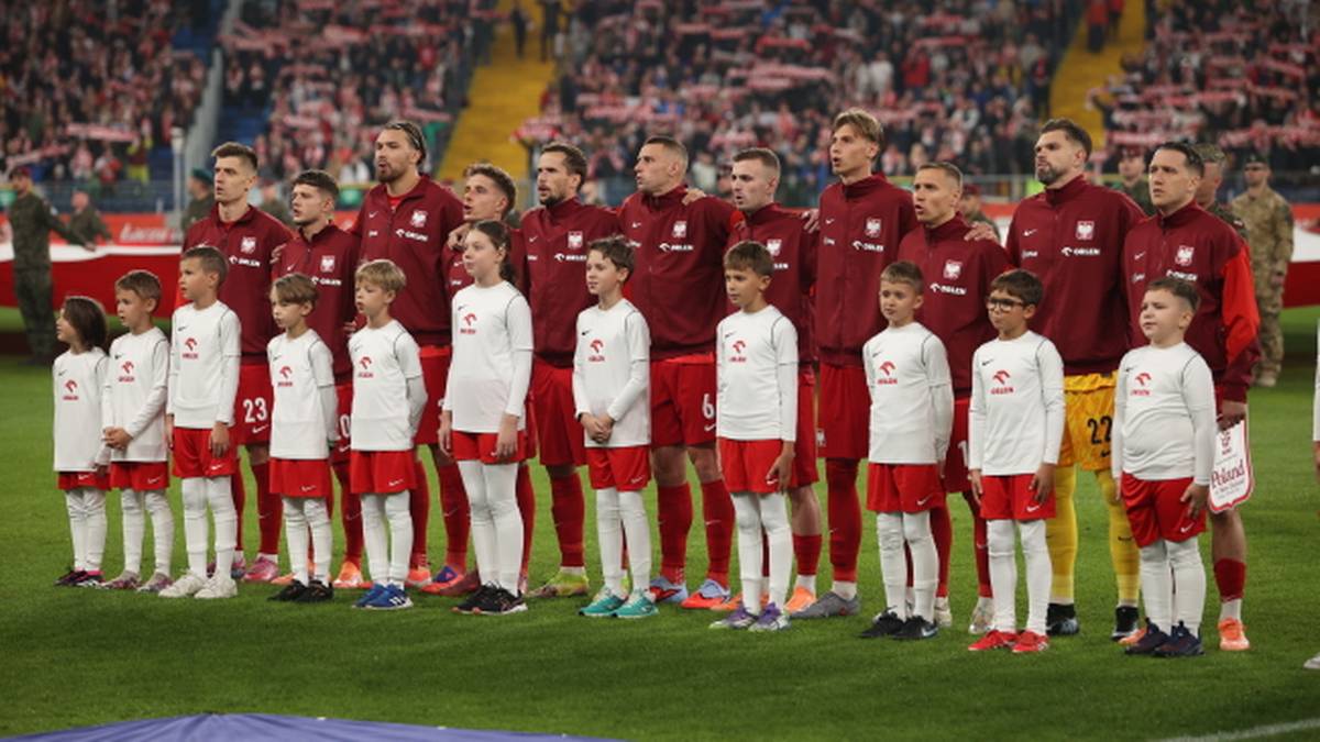 Grupa piłkarzy reprezentacji Polski w białych koszulkach stoi na boisku przed trybunami kibiców.