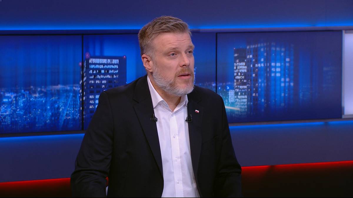 Mężczyzna w średnim wieku z brodą, ubrany w czarną marynarkę i białą koszulę, mówi patrząc w kamerę. Tło stanowią ekrany z widokiem nocnego miasta.