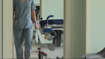 Osoba w niebieskim uniformie medycznym idzie w stronę łóżka szpitalnego.