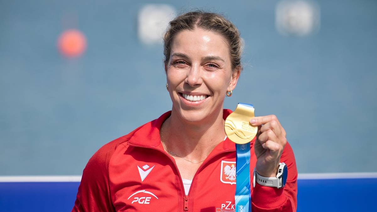 Kobieta w czerwonej bluzie sportowej trzyma złot medal na niebieskiej wstążce.