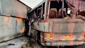 Uszkodzony autobus po wypadku, z widocznymi wybitymi oknami i zdeformowaną karoserią, stojący przy betonowej ścianie.