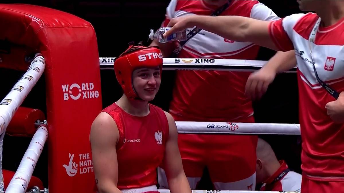 Młoda kobieta w czerwonym kasku bokserskim i stroju sportowym, uśmiecha się, stojąc w ringu.