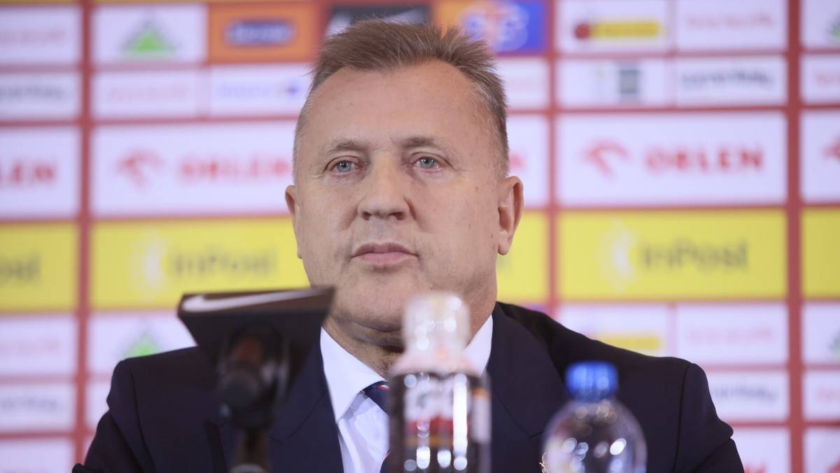 Mężczyzna w garniturze, prawdopodobnie trener piłkarski, siedzi za stołem podczas konferencji prasowej. Za nim znajduje się ściana z logotypami sponsor&oacute;w.