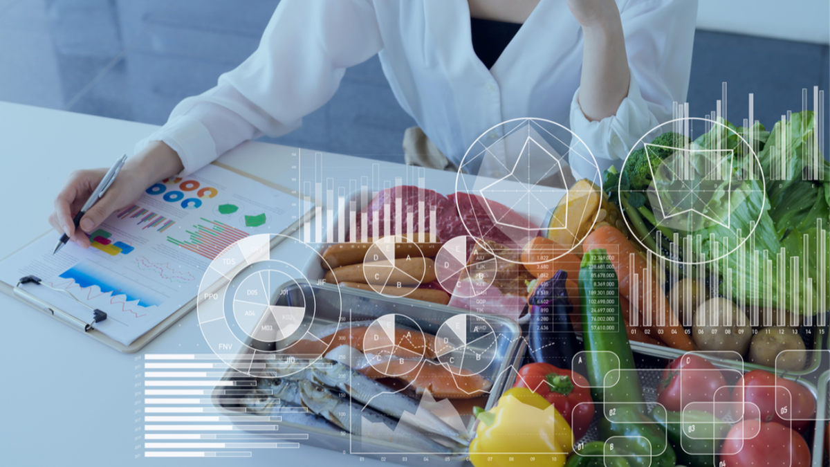 Kobieta analizująca wykresy przy biurku z r&oacute;żnorodnymi produktami spożywczymi, w tym warzywami, rybami i wędlinami.