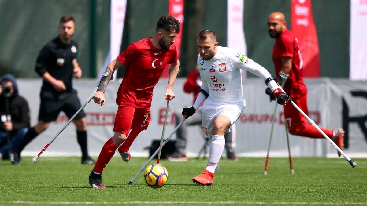 Dw&oacute;ch piłkarzy, jeden w czerwonej koszulce z godłem Turcji, drugi w białej koszulce z godłem Polski, grają w piłkę nożną, używając kul ortopedycznych.