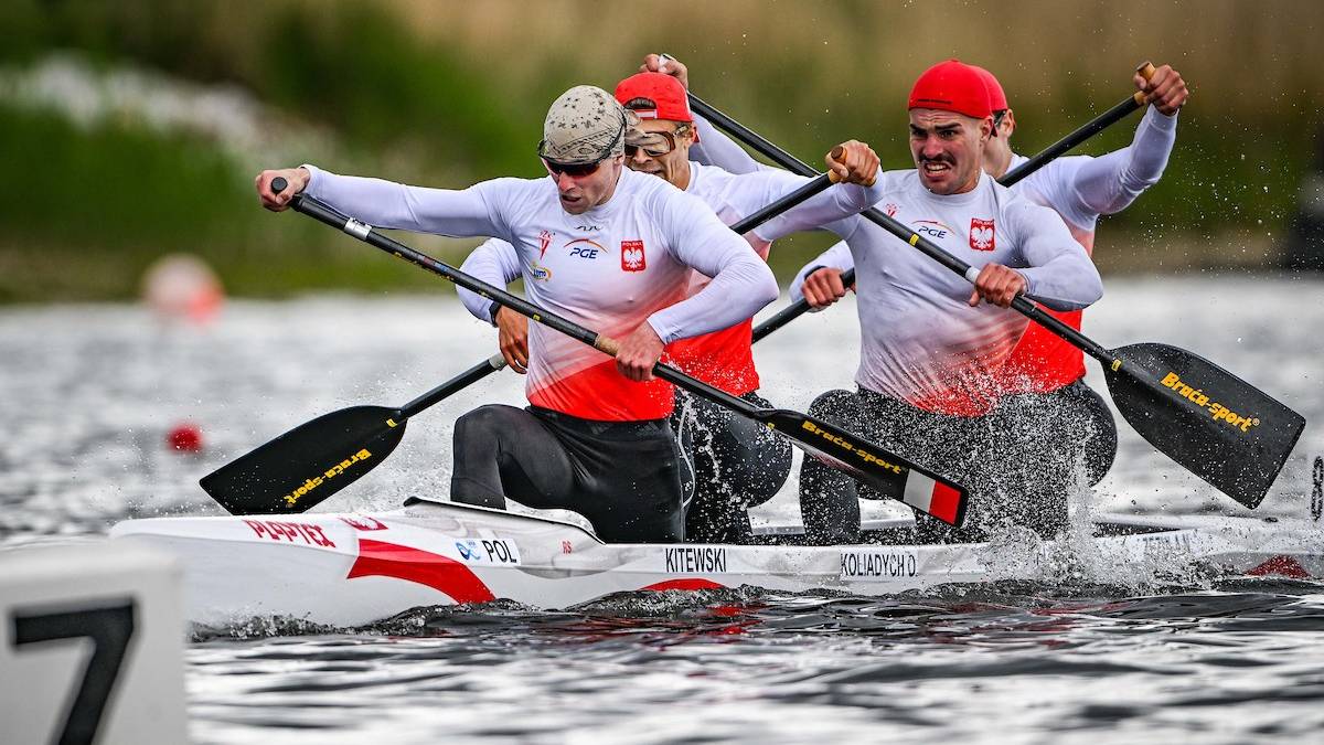 Czterech mężczyzn w kajaku z polskimi napisami, wiosłuje zgodnie z rytmem, przygotowując się do wyścigu.