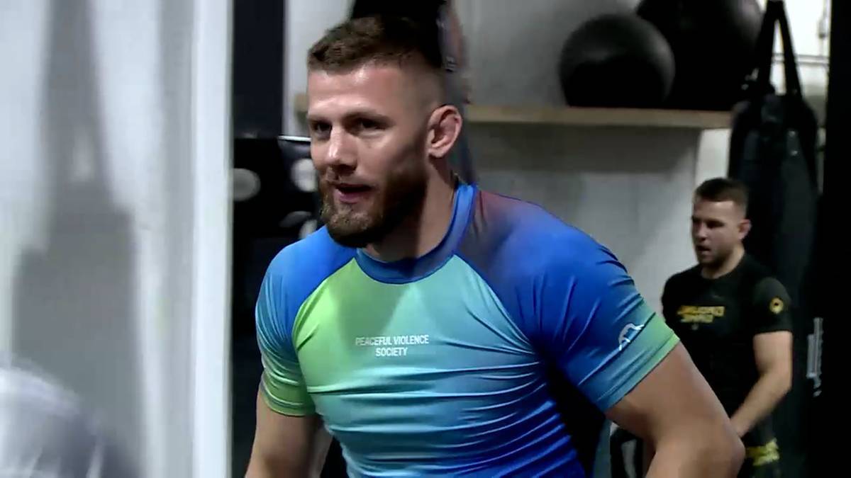 Mężczyzna w niebiesko-zielonej koszulce sportowej z krótkim rękawem, brodaty, patrzy w bok, lekko uśmiechnięty.