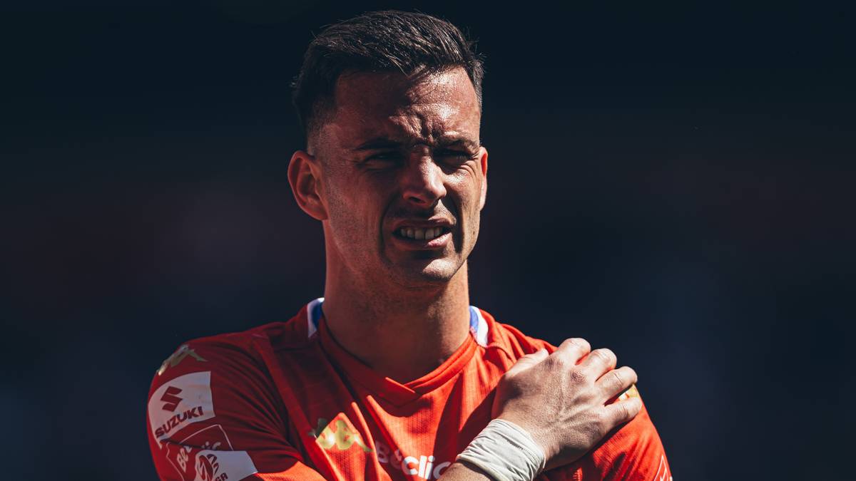 Młody piłkarz w czerwonej koszulce, trzymający się za lewy bark, z wykrzywioną twarzą wskazującą na b&oacute;l.