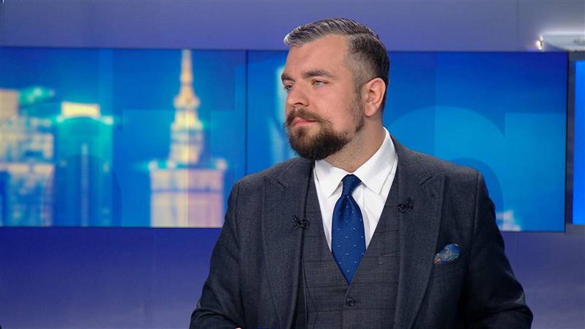 Mężczyzna w ciemnym garniturze i niebieskim krawacie, z zarostem, ubrany w kamizelkę, stoi przed niebieskim ekranem z widokiem na nocną panoramę miasta.