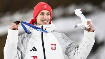 Mężczyzna w czerwonej czapce i białej kurtce z polskim godłem, trzyma medal na niebieskiej wstążce i pluszową zabawkę.