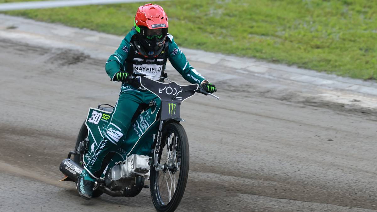 Żużlowiec w zielonym kombinezonie i czerwonym kasku jedzie na motocyklu po torze żużlowym.
