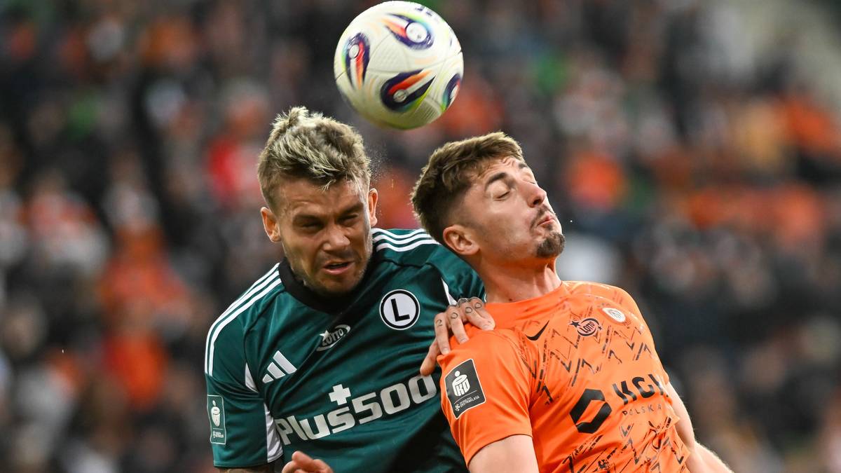 Dwóch piłkarzy walczy o piłkę w powietrzu podczas meczu piłki nożnej. Jeden z nich nosi zieloną koszulkę, a drugi pomarańczową.