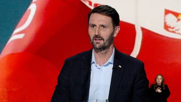 Mężczyzna w garniturze udziela wywiadu na tle polskiej flagi, z tłumaczką języka migowego w rogu kadru.