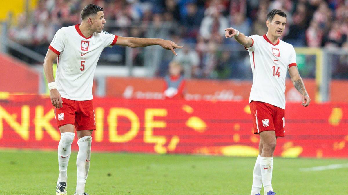 Dwóch polskich piłkarzy w białych koszulkach i czerwonych spodenkach, jeden wskazuje palcem w kierunku boiska, drugi patrzy w tym samym kierunku.