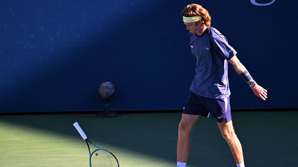 Gracz tenisa Andrej Rublev w niebieskim stroju i z żółtą opaską na głowie, stojący na korcie tenisowym z rakietą w ręku. Jego mina wyraża frustrację, a piłka leży niedaleko niego.