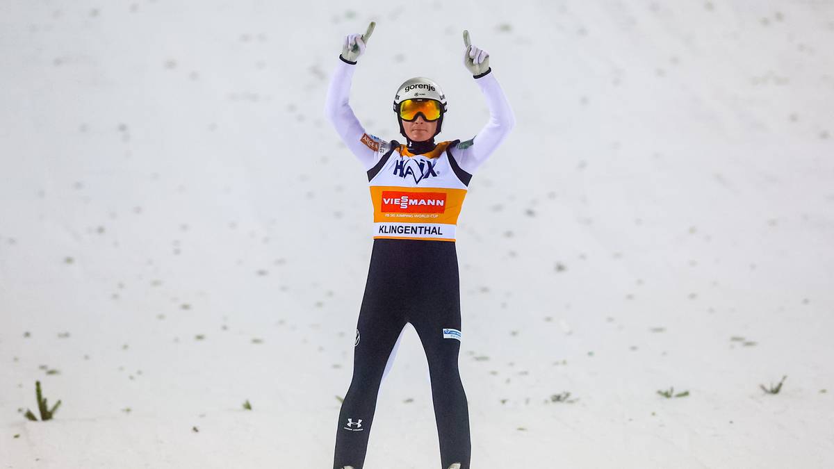 Skoczek narciarski w stroju, z nartami na nogach, unosi ręce w geście triumfu.