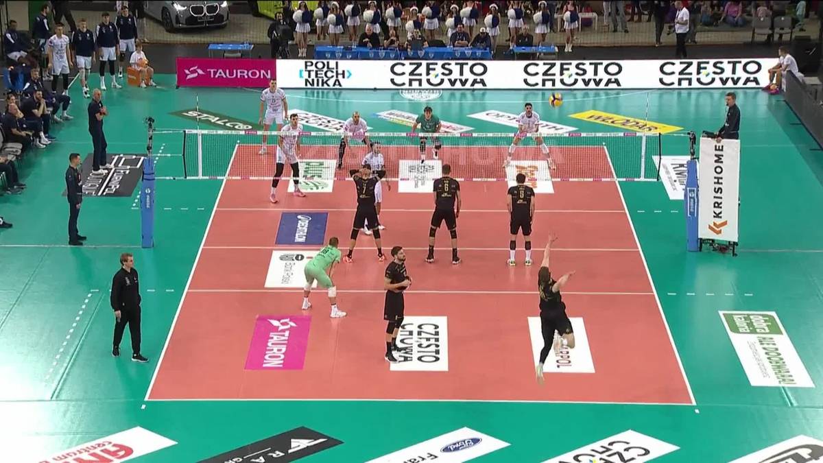 Drużyny siatkarskie rozgrywają mecz na hali sportowej. Zawodnicy w czarnych i żółtych strojach rywalizują na boisku z czerwonym polem. W tle widać publiczność.