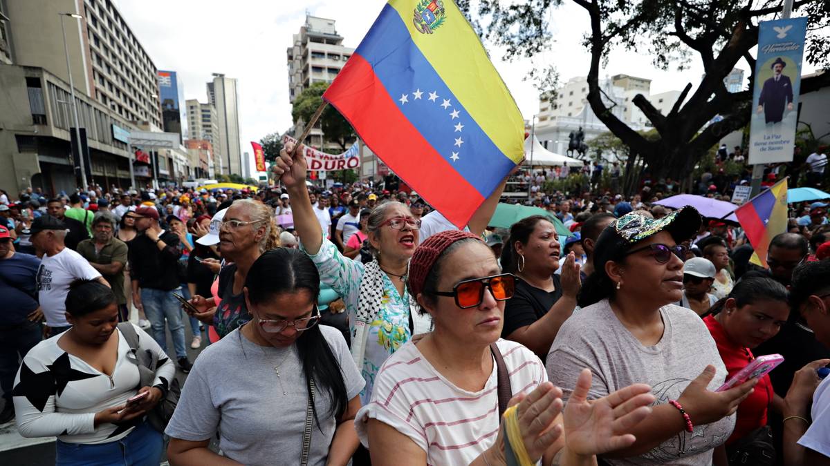 Grupa ludzi na ulicy trzyma w g&oacute;rze flagę Wenezueli. Niekt&oacute;rzy klaszczą i krzyczą.