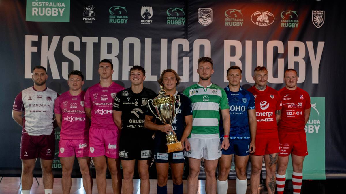 Dziewięciu młodych mężczyzn w strojach sportowych pozuje do zdjęcia na tle banera "EKSTRALIGA RUGBY". Jeden z nich trzyma w rękach wielkie, złote trofeum.