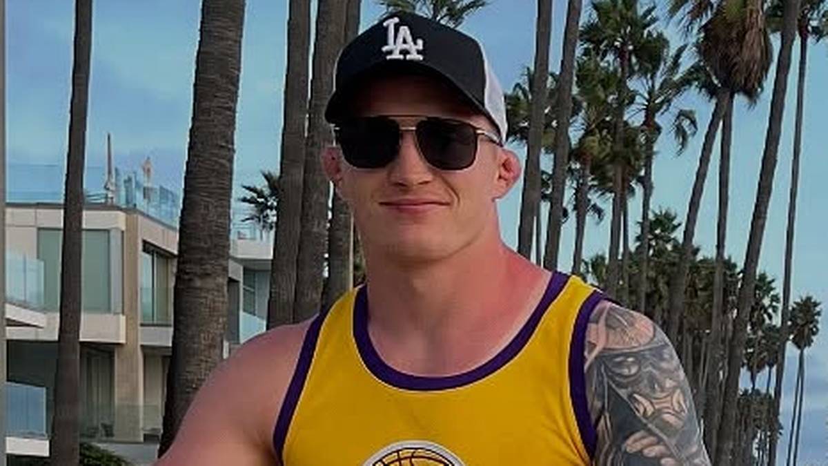 Uśmiechnięty mężczyzna w okularach przeciwsłonecznych i czapce z daszkiem "LA", ubrany w ż&oacute;łtą koszulkę bez rękaw&oacute;w, na tle palm i budynk&oacute;w.