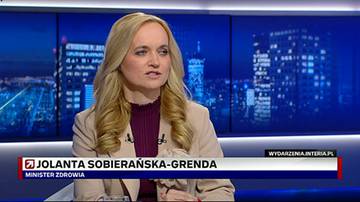 Kobieta z blond włosami i w beżowym żakiecie siedzi za biurkiem, m&oacute;wiąc do kamery. W tle widoczne są nocne panoramy miast na tle niebieskiego ekranu.