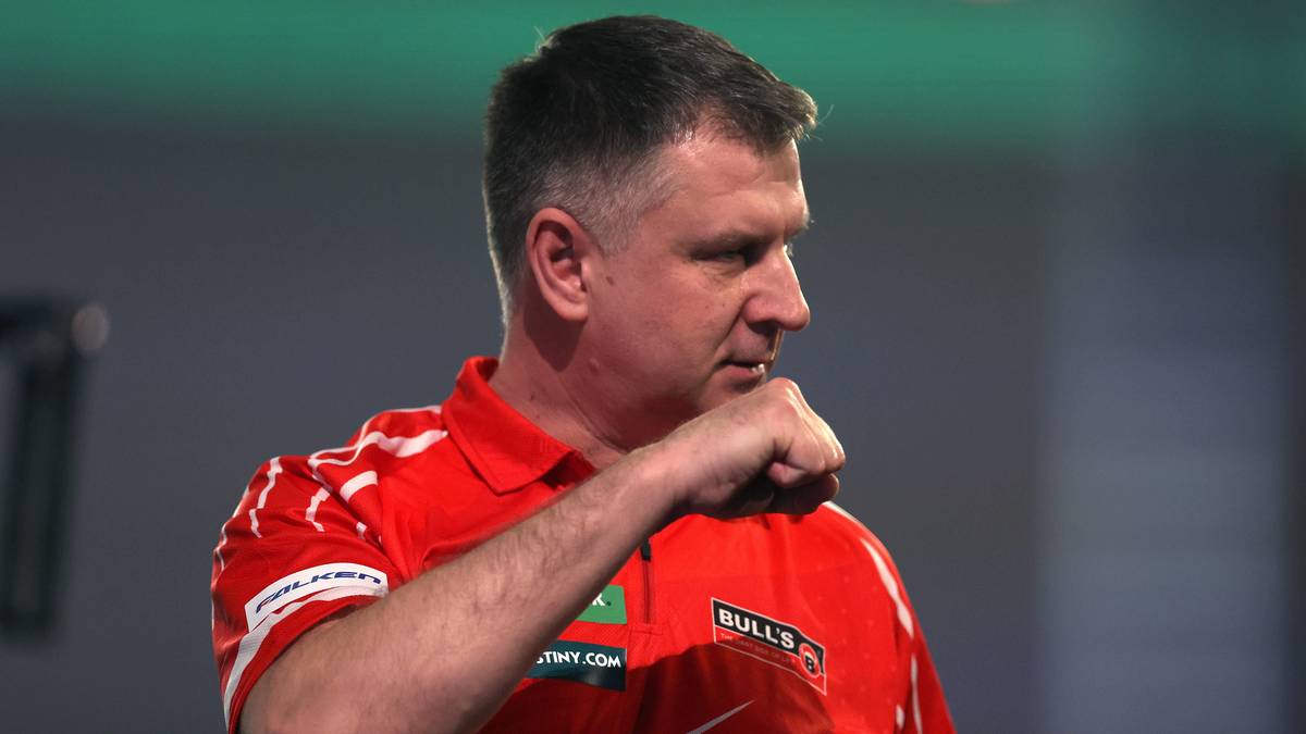 Krzysztof Ratajski w czerwonej koszulce z białym wzorem, patrzący w bok z zaciśniętą pięścią.