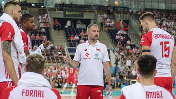 Trener siatk&oacute;wki zwraca się do swoich zawodnik&oacute;w w białych strojach z czerwonymi akcentami, stojąc na boisku w otoczeniu publiczności na trybunach.