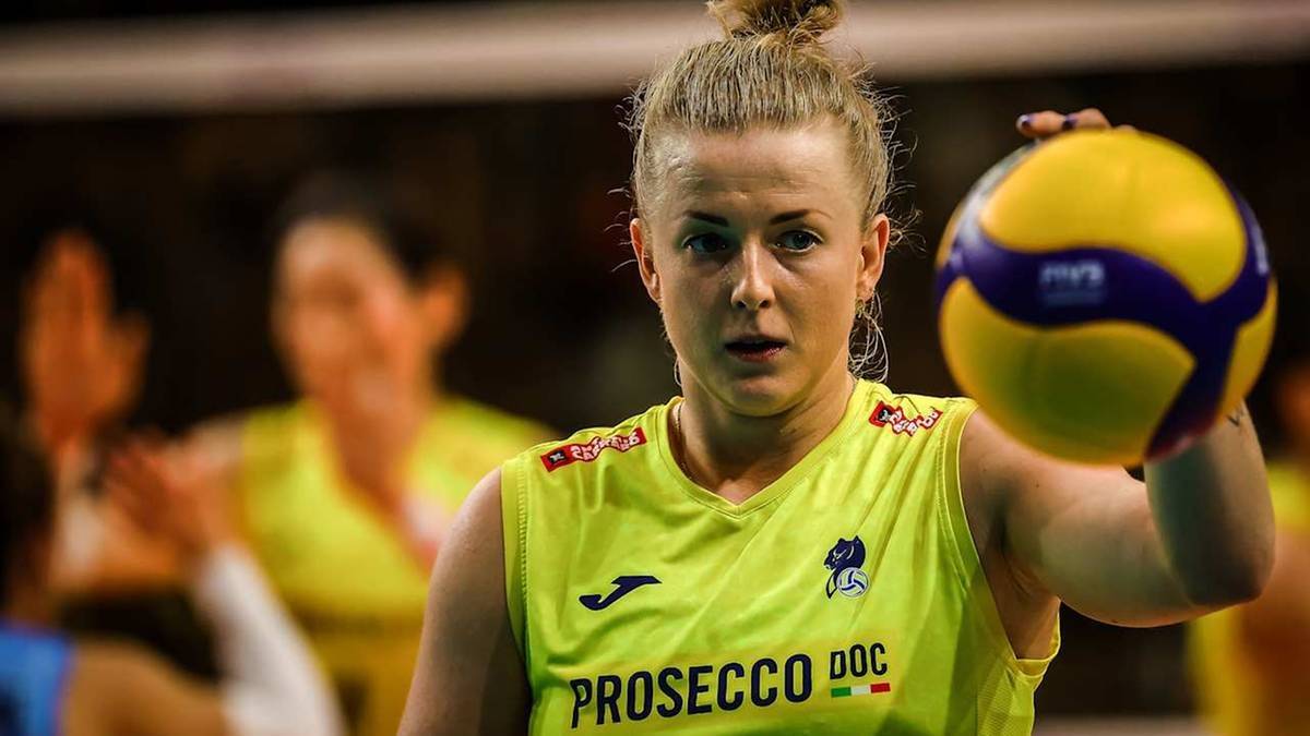 Siatkarka w żółtej koszulce z napisem "Prosecco Doc" trzyma piłkę do siatkówki.