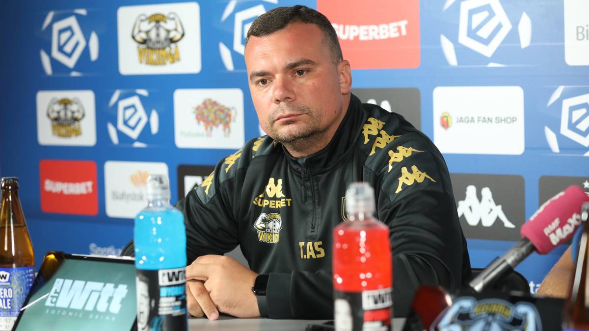 Mężczyzna w czarnej bluzie z logo Jagiellonii siedzi przy stole podczas konferencji prasowej. W tle widoczne są logo i napisy.