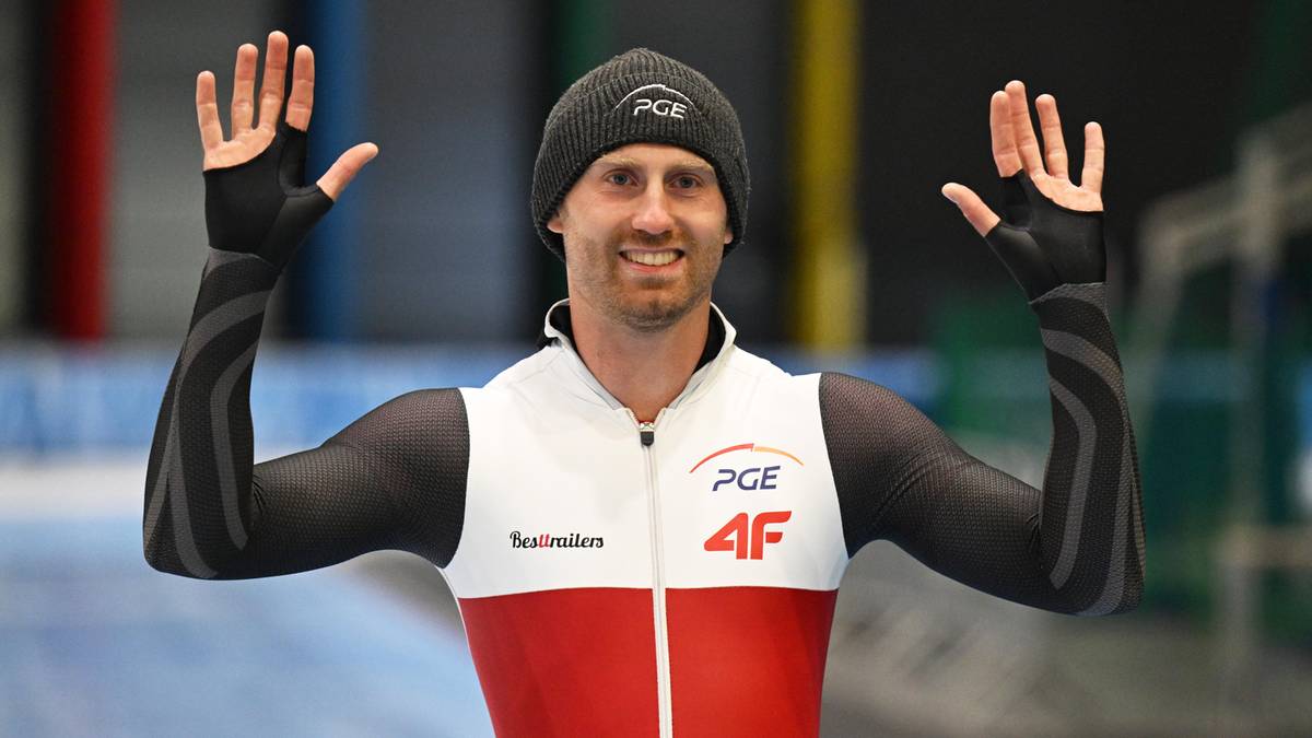 Łyżwiarz szybki w czapce i stroju sportowym z uniesionymi rękami, uśmiecha się.