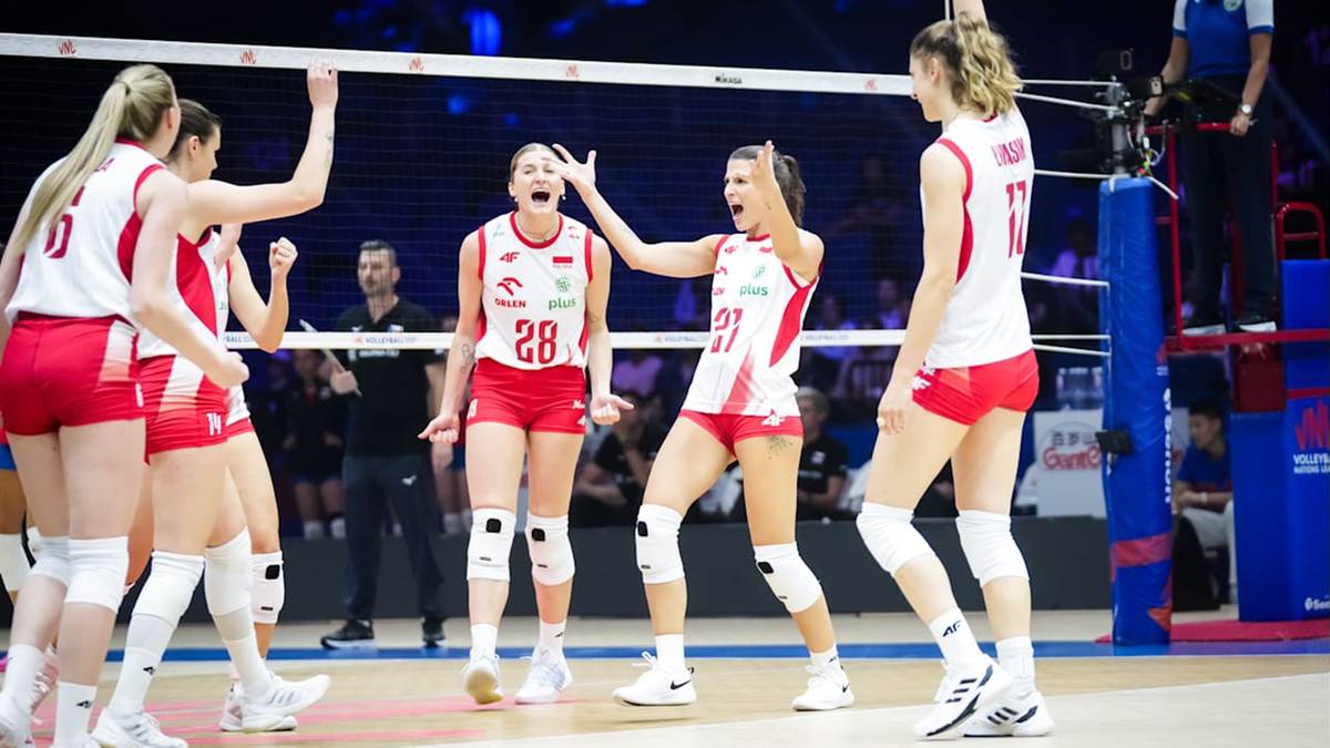 Grupa siatkarek w białych strojach z czerwonymi akcentami, świętujących na boisku do siatkówki.