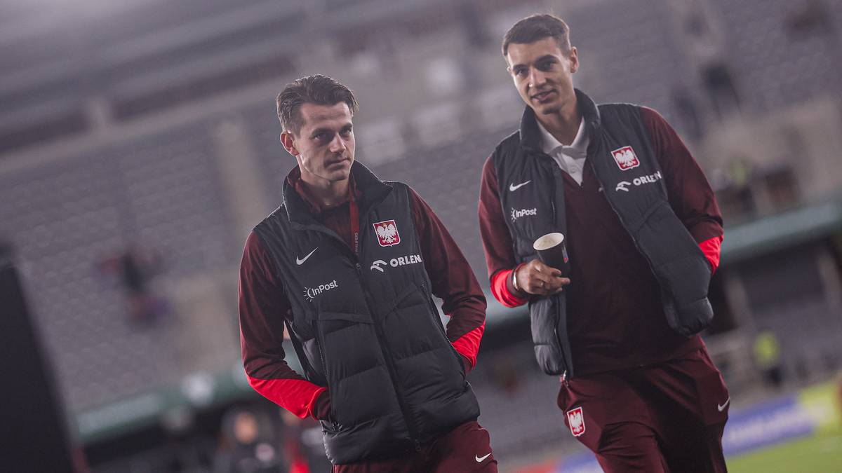 Dw&oacute;ch mężczyzn w kamizelkach reprezentacji Polski stoi na boisku piłkarskim, jeden z nich trzyma kubek.