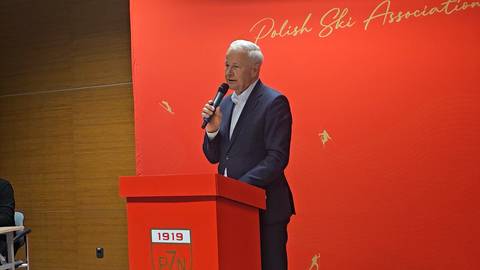 Apoloniusz Tajner przemawia do mikrofonu stojąc za czerwona m&oacute;wnicą z napisem 1919 na tle czerwonej ściany z logo Polish Ski Association.