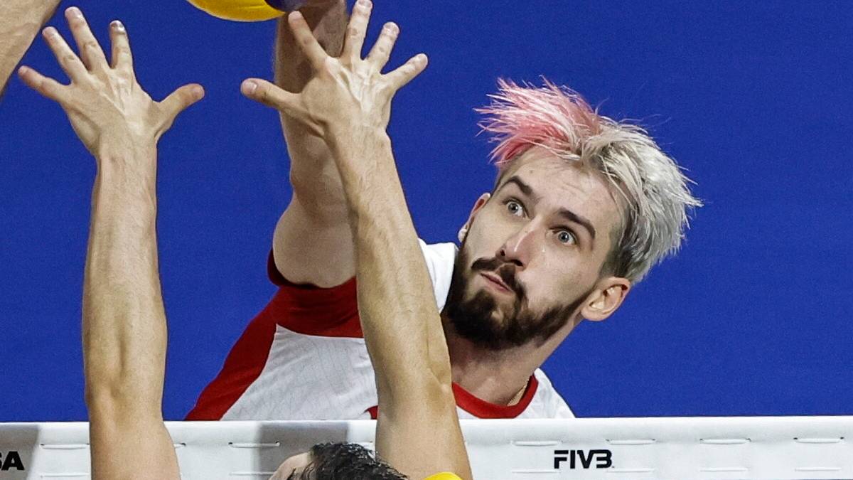 Siatkarz z różowymi pasemkami we włosach wykonuje wysoki wyskok do ataku, jego dłonie są wyciągnięte w kierunku piłki nad siatką. Naprzeciw niego drugi zawodnik próbuje zablokować.