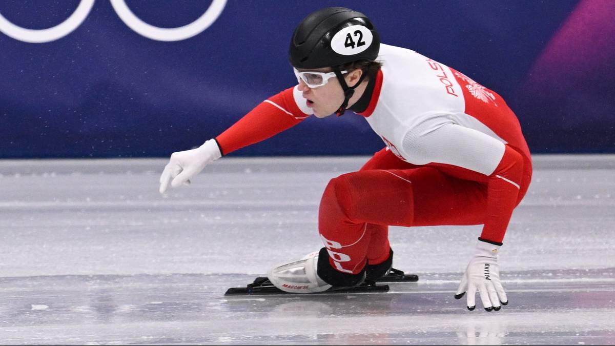 Zawodniczka w kasku i kombinezonie na lodowisku podczas zawodów short track, z symbolem kół olimpijskich w tle.