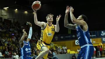 Trzech koszykarzy podczas meczu. Jeden gracz w ż&oacute;łtym stroju z numerem 33 na plecach wykonuje rzut do kosza, podczas gdy dwaj przeciwnicy w niebieskich strojach pr&oacute;bują go zablokować.