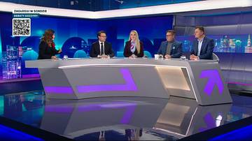 Cztery osoby siedzą przy futurystycznym stole w studiu telewizyjnym. Jedna kobieta stoi po lewej stronie, a trzy osoby siedzą za stołem.