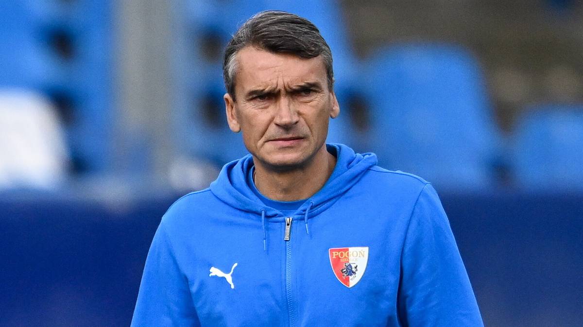 Trener na boisku piłkarskim, w tle piłka i pachołek.