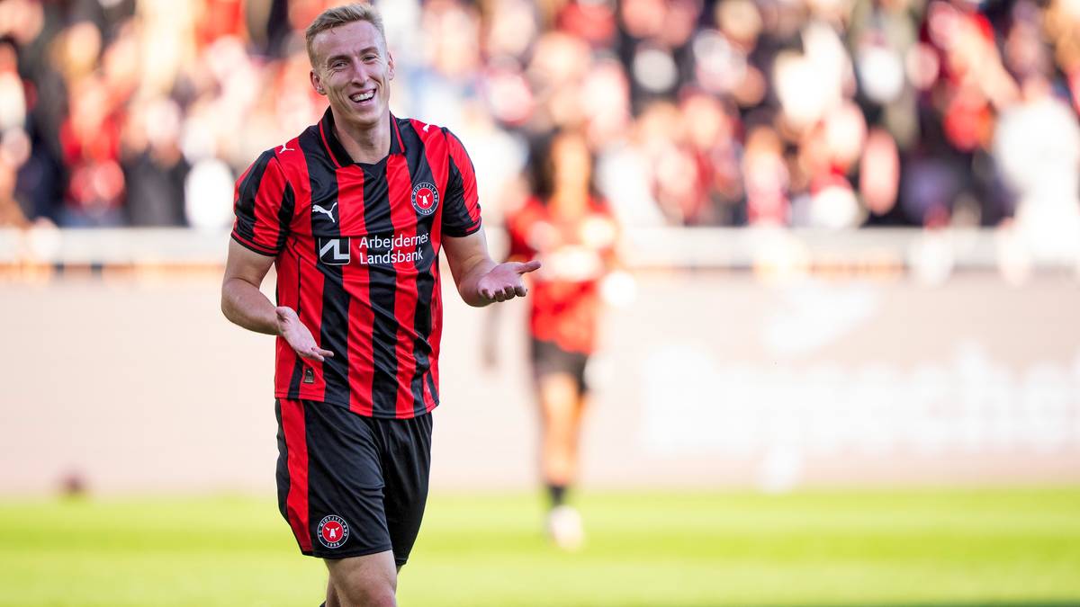 Piłkarz w czerwono-czarnej koszulce z pionowymi pasami uśmiecha się i gestykuluje rękami, radośnie celebrując udany strzał lub bramkę.