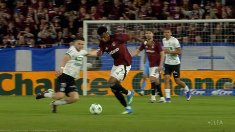 Dw&oacute;ch piłkarzy na boisku, jeden w białej koszulce, drugi w bordowej, walczy o piłkę. W tle trybuny z widzami.