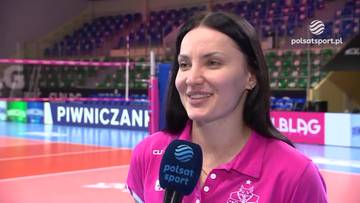 Kobieta w r&oacute;żowej koszulce z mikrofonem w dłoni, uśmiechnięta, na tle hali sportowej z boiskiem do siatk&oacute;wki.