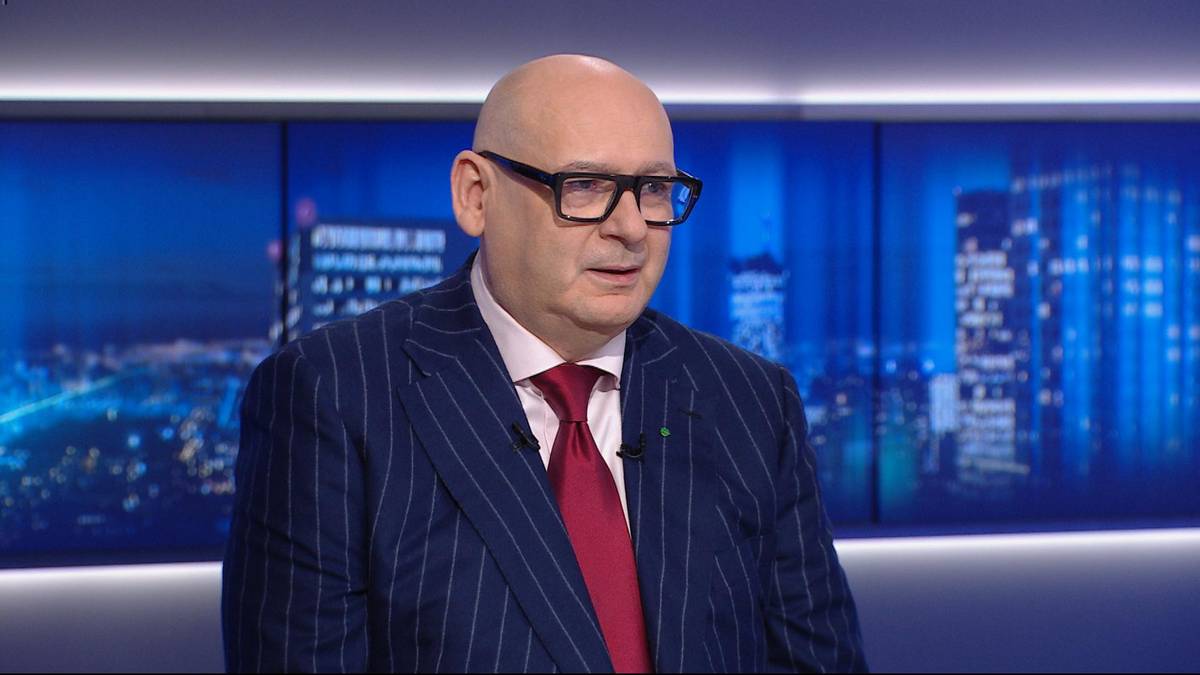 Mężczyzna w okularach i granatowym garniturze w paski z czerwonym krawatem stoi przed niebieskim ekranem z widokiem nocnego miasta.