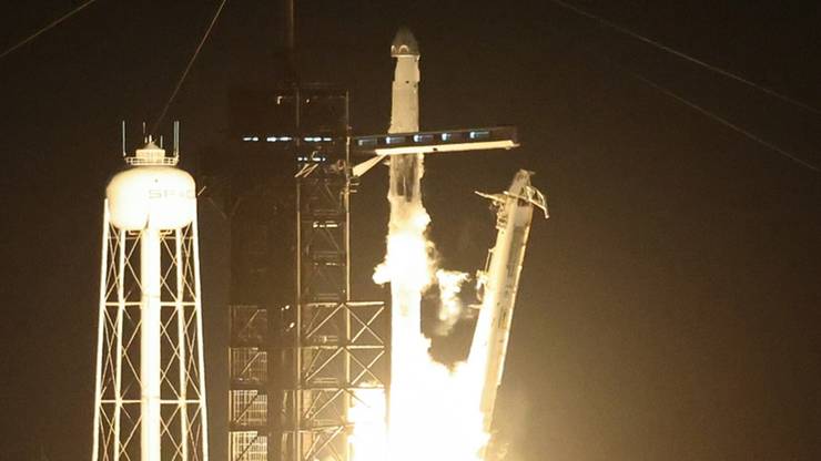 Rakieta kosmiczna startująca w nocy, z jasnymi płomieniami wydobywającymi się z silnik&oacute;w. Obok znajduje się wieża startowa i zbiornik z logo SpaceX.